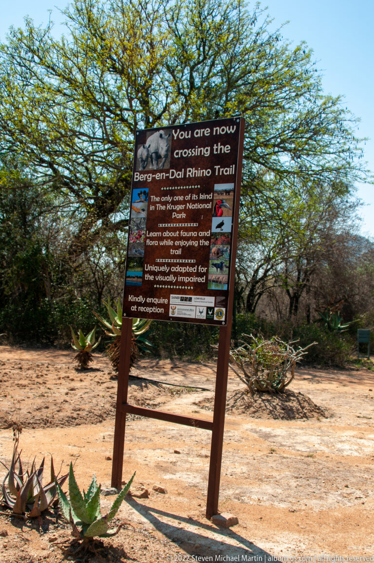 Wooden trail sign in dry bush announcing a crossing on the Berg-en-Dal Rhino Trail and listing rules.