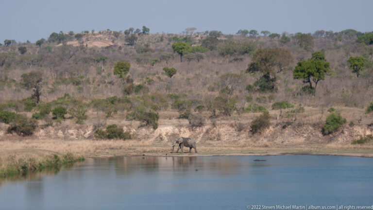 Lone African elephant standing on the far bank of a wide waterhole below a dry wooded hillside.
