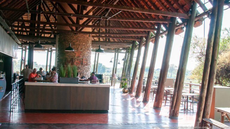 Open-air rest-camp pavilion with timber posts, high roof, service counter, tables, and several visitors.