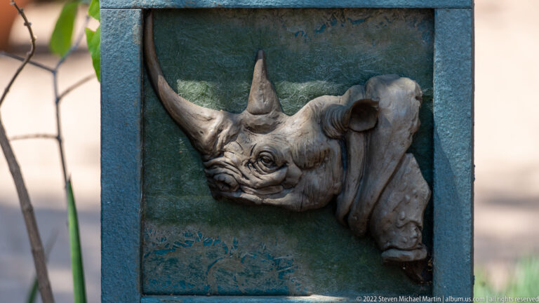 Gray rhinoceros head relief with two horns mounted on a weathered green rectangular plaque.
