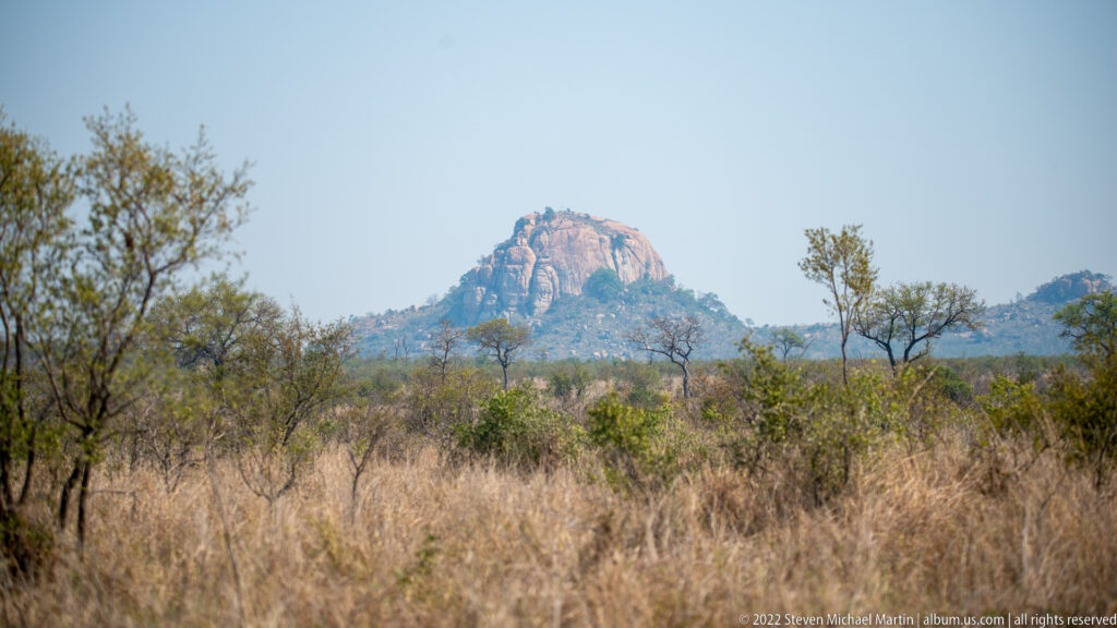 Rounded granite kopje rising above dry grass and scattered bushveld trees beneath a clear blue sky.