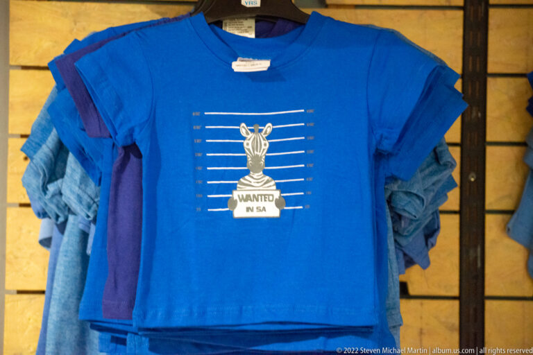 Blue souvenir T-shirt hanging on display with a zebra graphic behind bars and the words 'WANTED IN SA.'