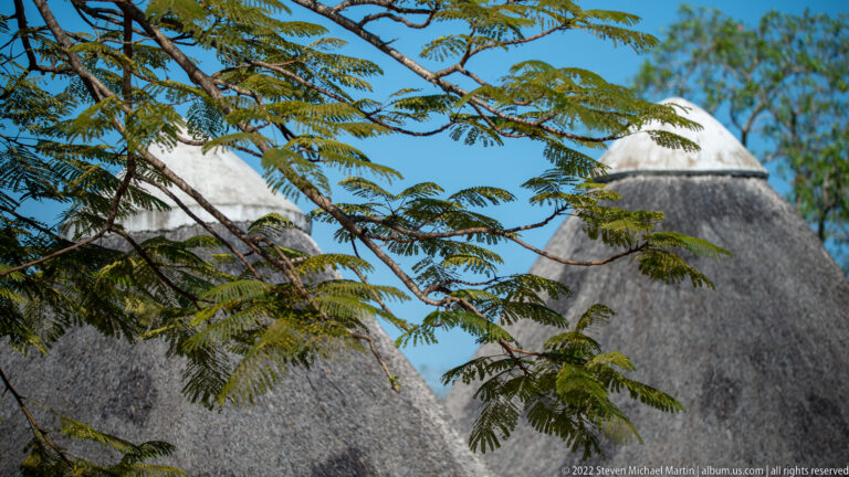 Pointed conical thatched roofs seen through leafy branches against a clear blue sky at a Kruger visitor facility.