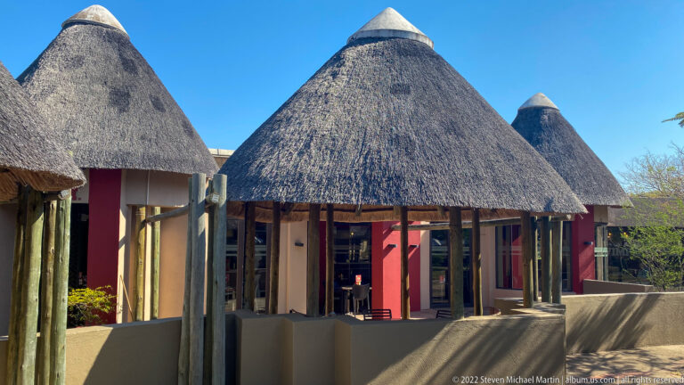 Visitor building with multiple conical thatched roofs, timber posts and red wall sections under a clear blue sky.