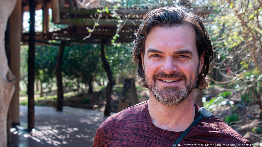 Close portrait of a smiling man outdoors with a shaded timber walkway and trees behind him.