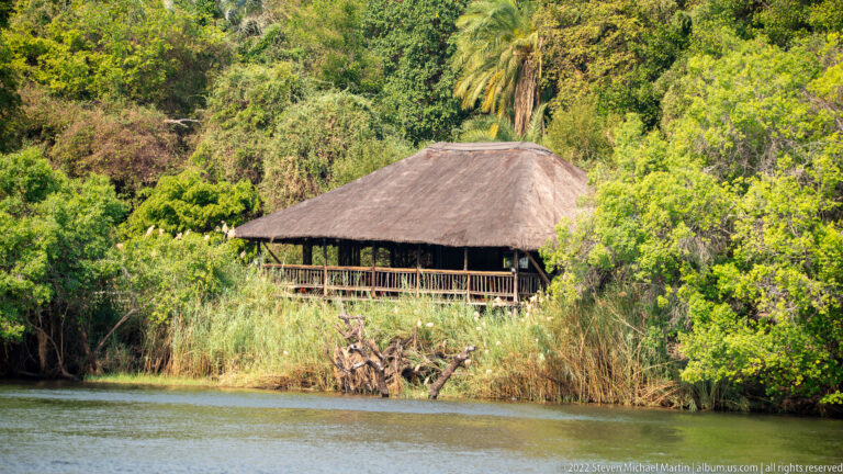 Thatched riverside lodge partly hidden by dense green trees and reeds above calm water.