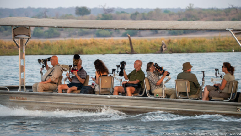 Open safari boat carrying seated wildlife photographers with long lenses across the river.