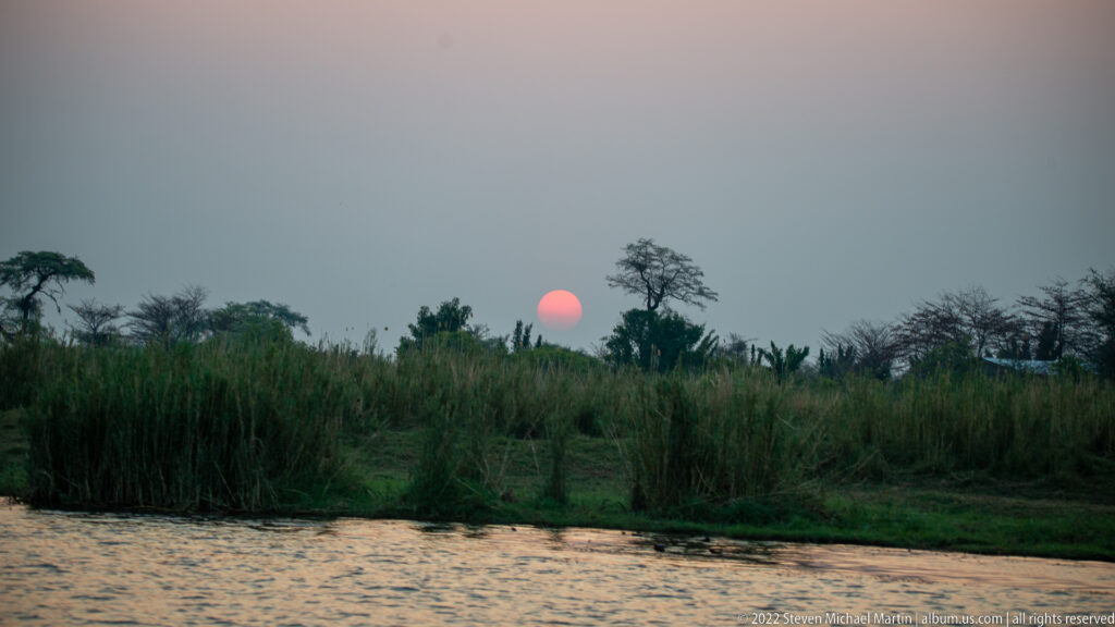 Pink sun setting behind tall reeds and scattered trees across a calm river.