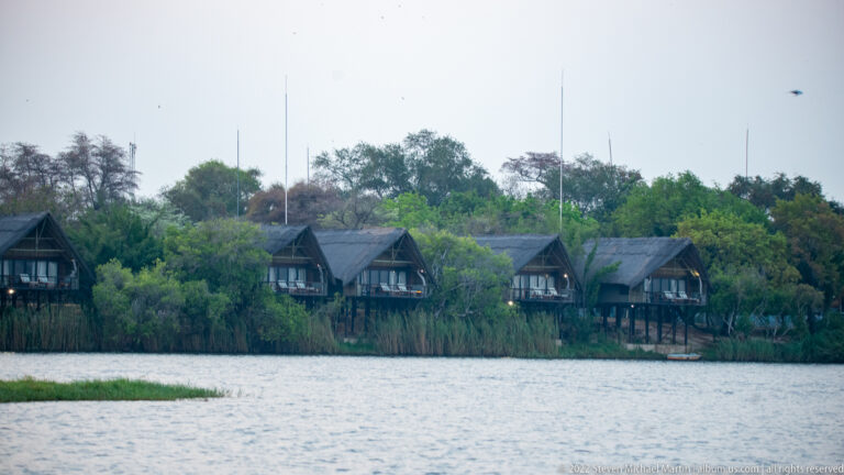 Row of thatched-roof lodge chalets on a green riverbank, seen across the water.