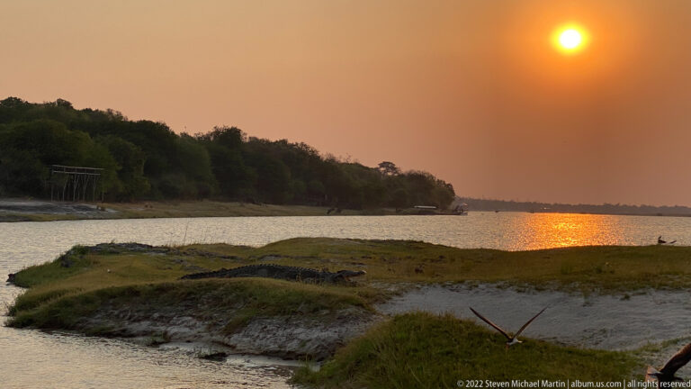 Orange sun over a broad river bend with bright reflection, low islands, and dark wooded shoreline.