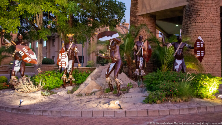 Life-size decorative figures with spears and patterned shields arranged among rocks and plants outside a resort building.