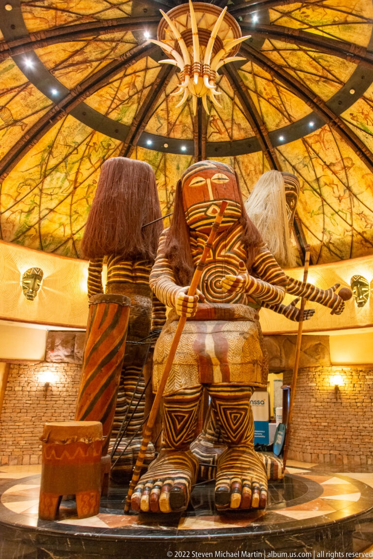 Large stylized carved figures with painted geometric patterns and fiber hair beneath a radial thatched ceiling.