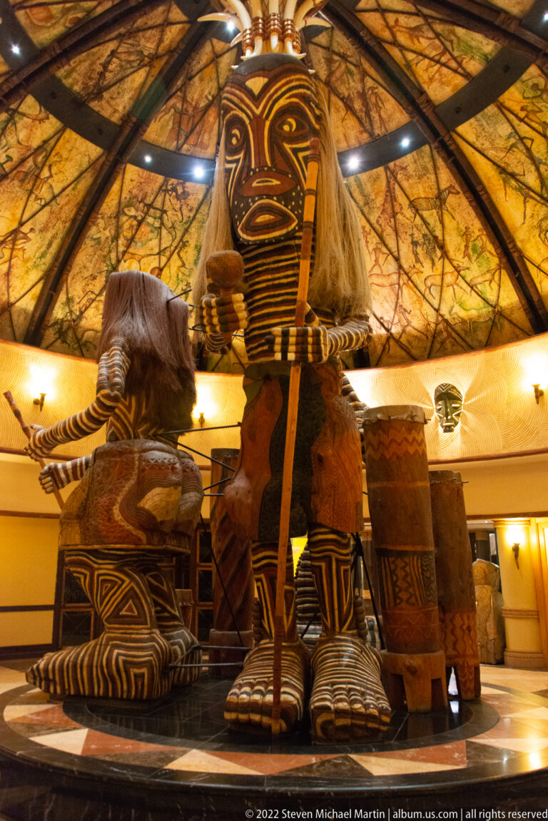 Low-angle view of a towering patterned carved figure with large feet and fiber hair inside a circular lobby.