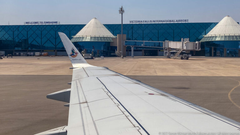 Aircraft wing facing the blue-glass terminal labeled Victoria Falls International Airport, with white conical roof structures.