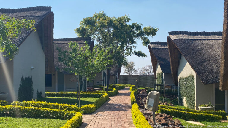Brick walkway between white lodge buildings with steep thatched roofs, clipped hedges, and green lawns.