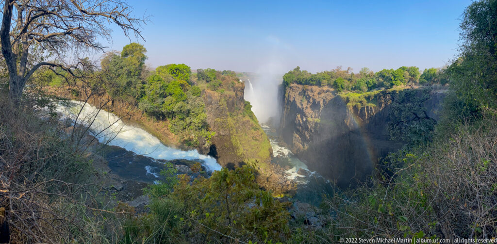 Wide view of Victoria Falls with several waterfall curtains, rocky gorge, mist, rainbow, and foreground branches.