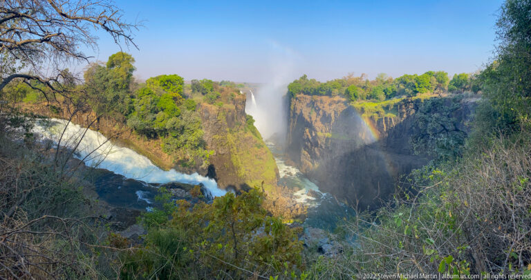 Vegetation-framed view of Victoria Falls, deep gorge, rushing water, mist, and a central rainbow.