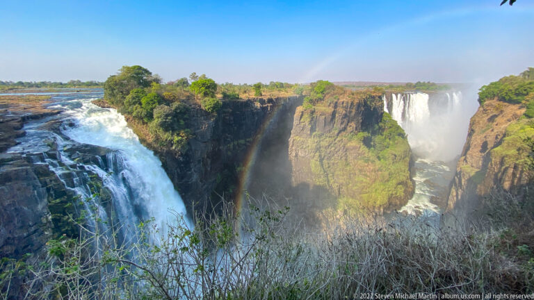 Zambezi River spilling over the near edge of Victoria Falls with a broad opposite waterfall and rainbow in the gorge.