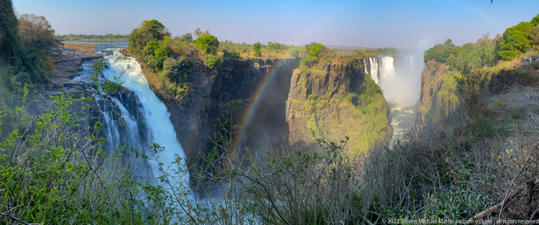 Near waterfall plunging beside a vivid rainbow, with another waterfall across the basalt gorge and shrubs in foreground.