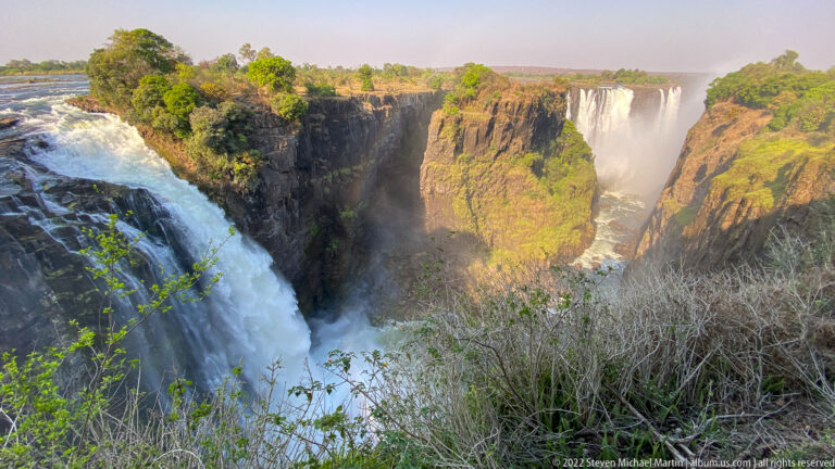 White water pouring over the near brink of Victoria Falls beside sunlit basalt cliffs and a distant waterfall.
