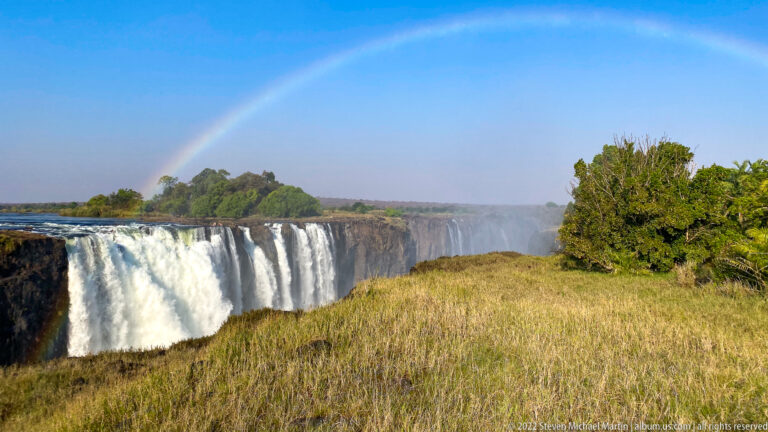 Broad curtain of Victoria Falls beside a dry grassy bank with trees, mist, and a faint rainbow.