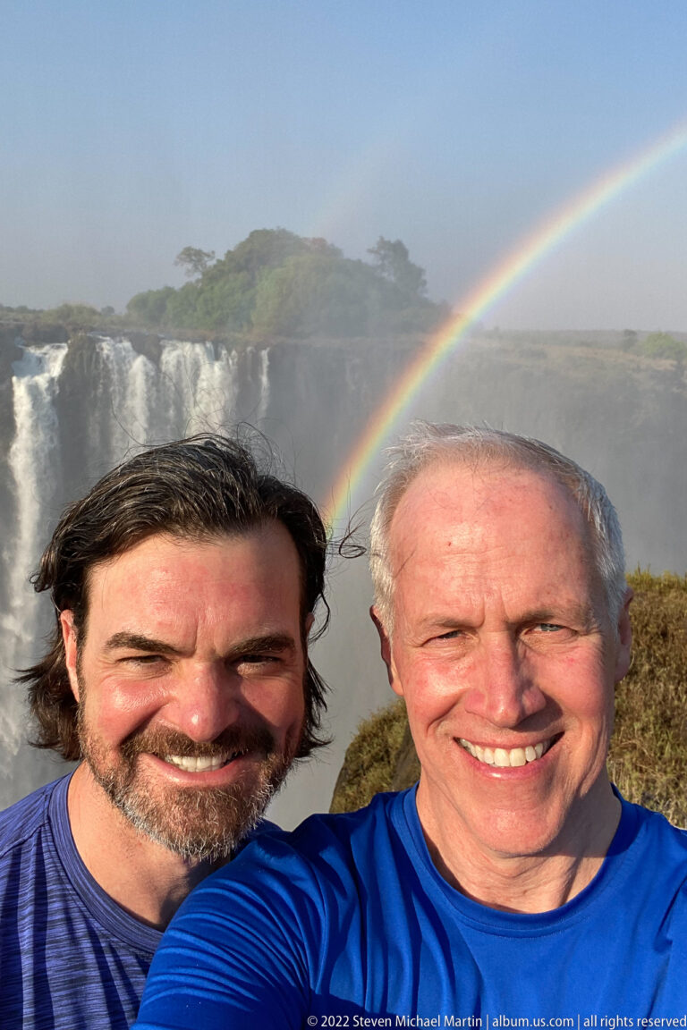 Two smiling visitors taking a selfie with Victoria Falls and a rainbow behind them.