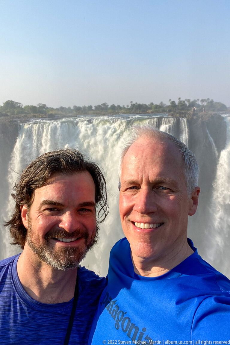 Two smiling visitors in close foreground with the white curtain of Victoria Falls directly behind them.