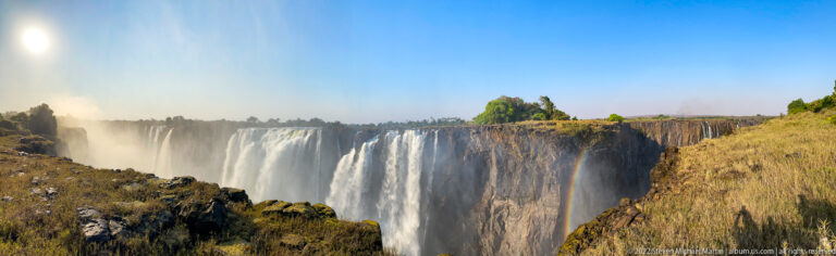 Long oblique view of Victoria Falls dropping over a dark cliff beside dry grass and rising mist.