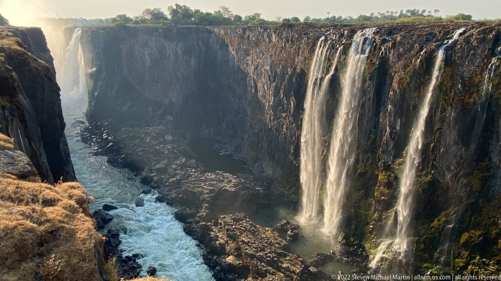Several narrow waterfalls descending a tall dark basalt cliff into a rocky, fast-flowing gorge.