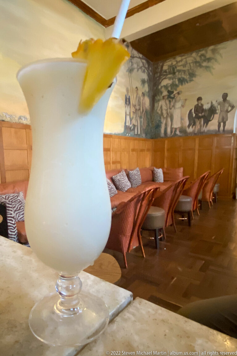 Pale frozen cocktail in a stemmed glass with citrus garnish, with lounge seating and a mural blurred behind.