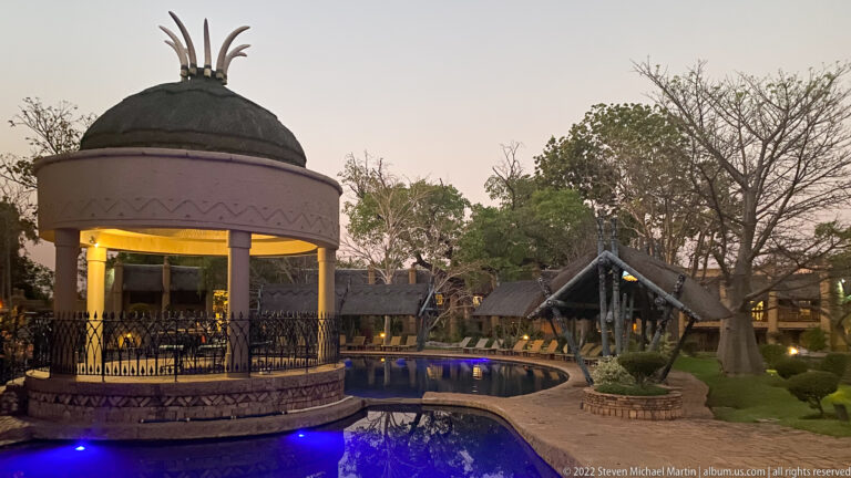 Domed open pavilion beside a curving blue-lit swimming pool, with thatched buildings and landscaped grounds at dusk.