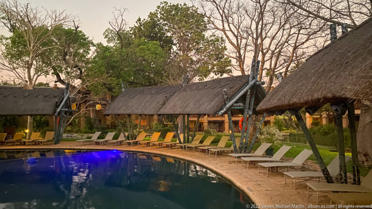 Curving blue swimming pool at dusk beside thatched lodge buildings, verandas, trees, and rows of lounge chairs.