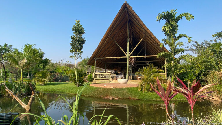 Open-sided pavilion with a steep thatched roof beyond a landscaped pond, palms, reeds, and colorful foliage.