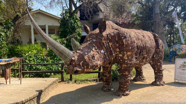 Life-size rhinoceros sculpture made from overlapping rust-brown metal pieces, standing outdoors among garden vegetation.