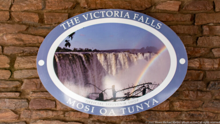 Round souvenir shop sign reading The Victoria Falls and Mosi Oa Tunya, with a waterfall and rainbow photograph.
