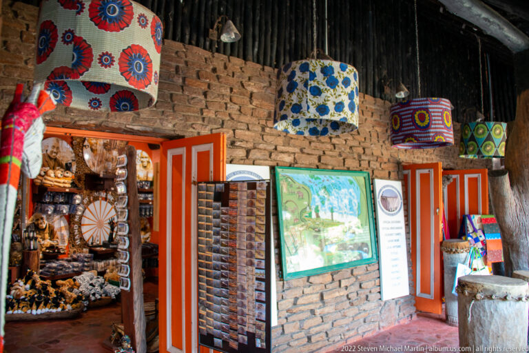 Souvenir shop interior with patterned hanging lamps, textiles, carved goods, display racks, brick walls, and an orange doorway.