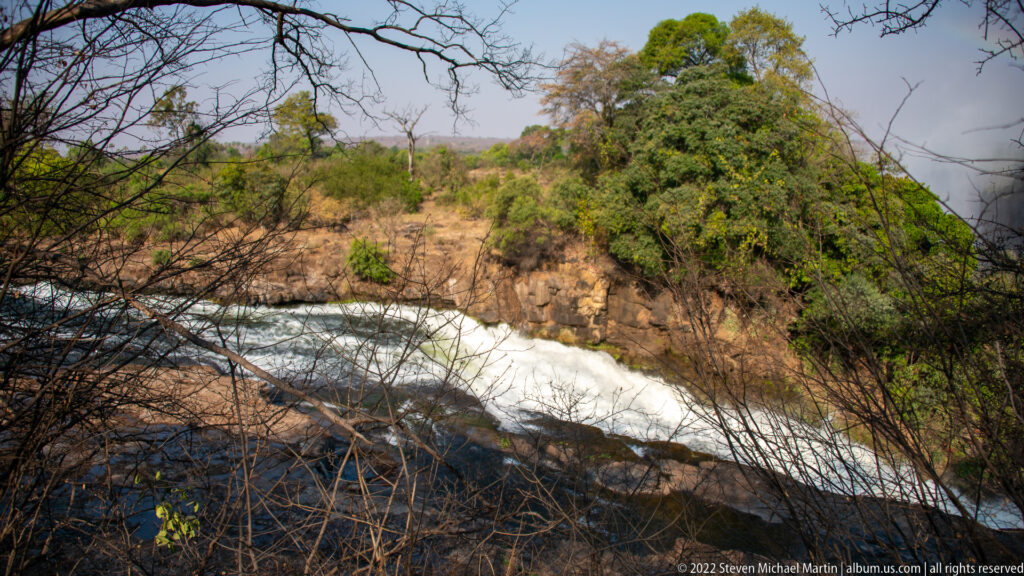White-water rapids flowing through a rocky Zambezi River channel bordered by woodland and basalt outcrops.