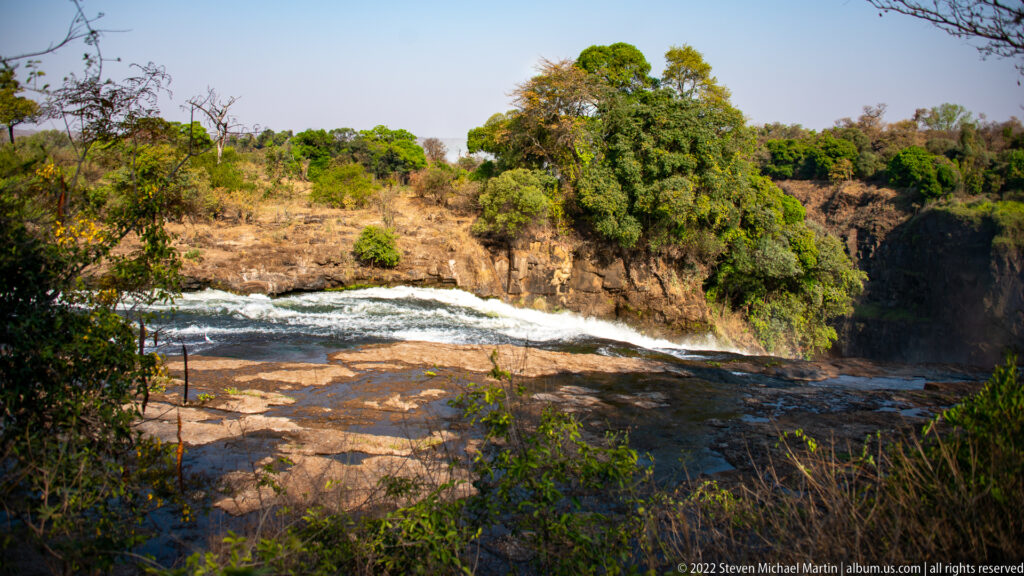 Broad rocky Zambezi River channel with low rapids, exposed shelves, wooded banks, and a drop toward the gorge.