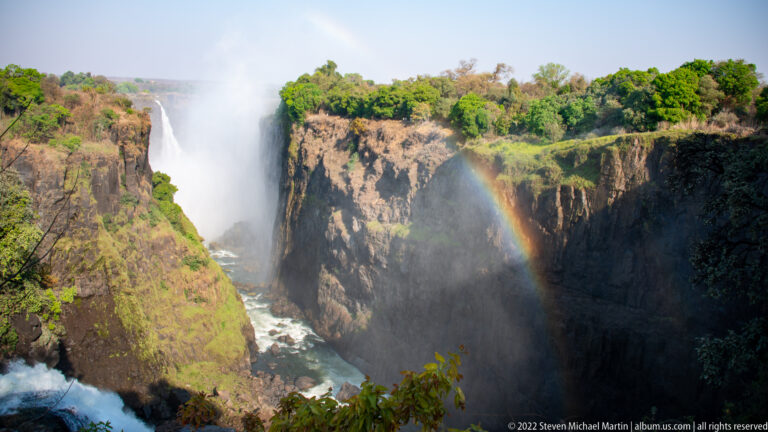Waterfall plunging between steep basalt cliffs with rising mist, green vegetation, and a bright rainbow in the gorge.