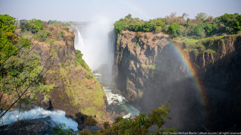 Closer view of a Victoria Falls cascade beside basalt cliffs, with a vivid rainbow arcing through heavy spray.