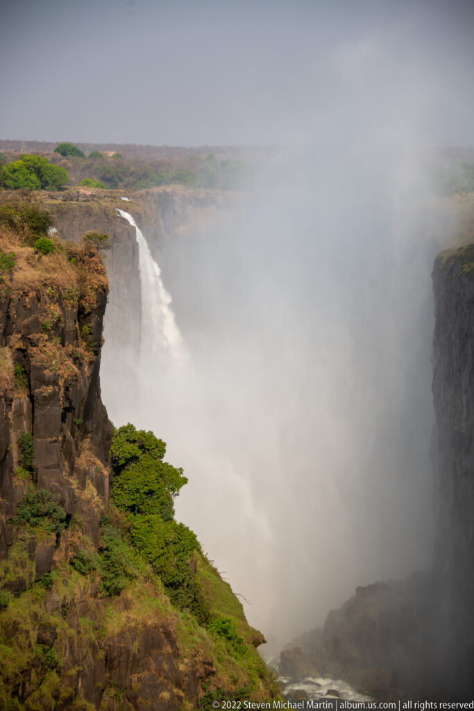 Basalt cliff rim and green vegetation beside a Victoria Falls cascade largely hidden by dense white mist.
