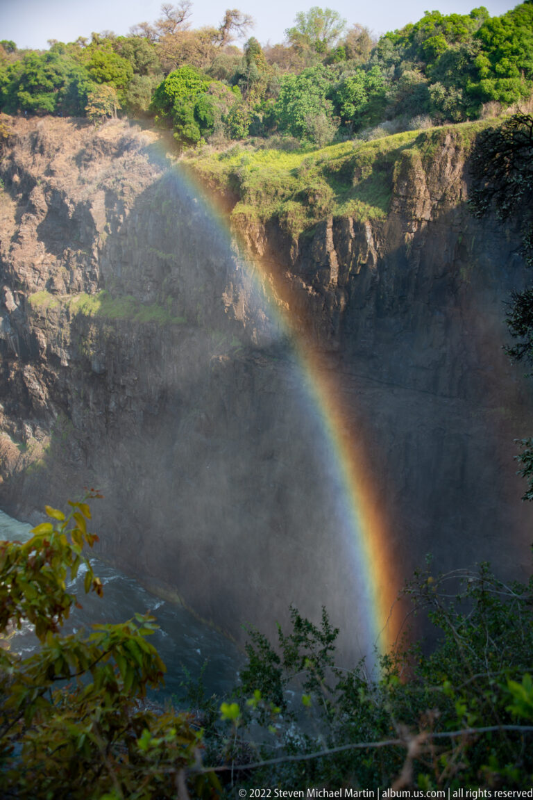 Broad rainbow descending through waterfall mist against a dark basalt cliff, framed by green foliage.