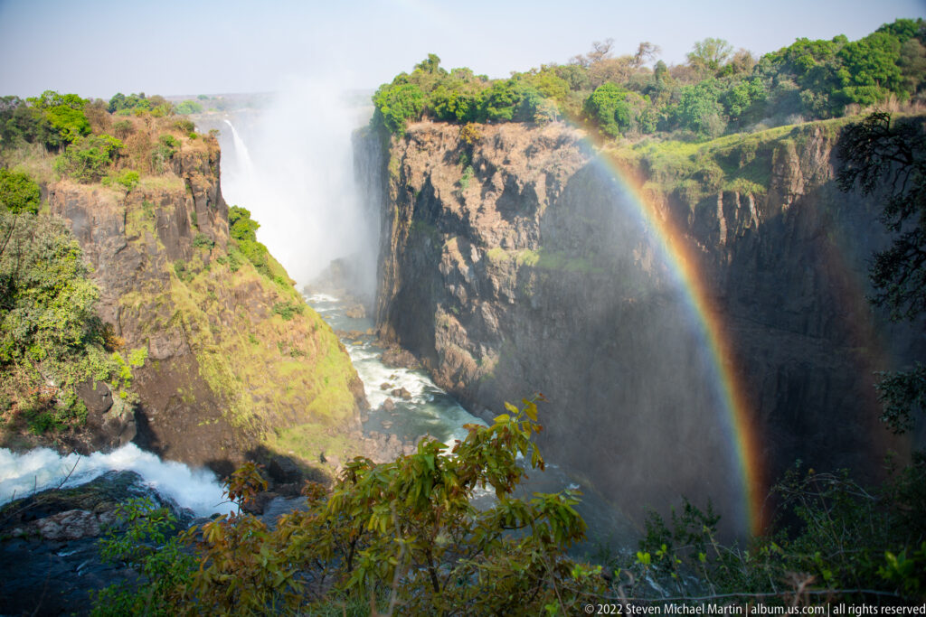 Victoria Falls cascade plunging into a basalt gorge beside a near-complete rainbow, with green vegetation on the rim.