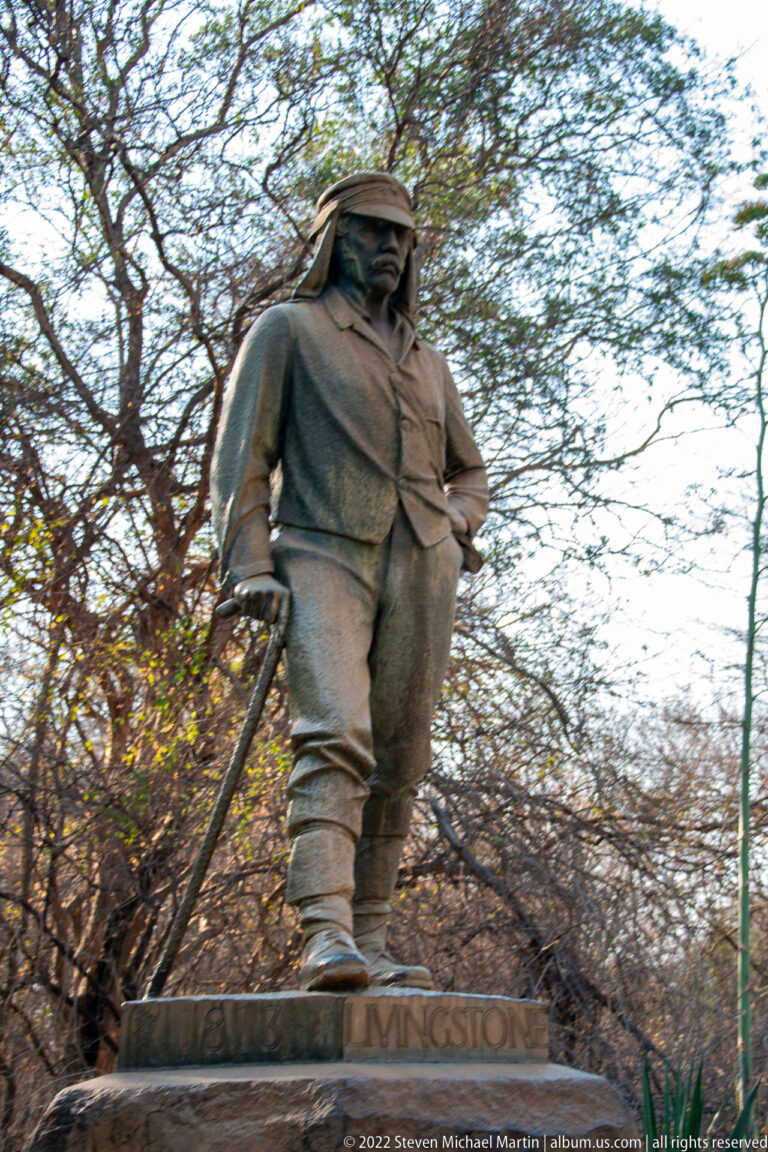 Full-length statue of David Livingstone in travel clothing holding a staff, standing on a plinth among trees.