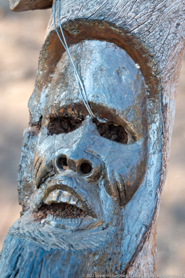 Close view of a weathered wooden carving with a human face, heavy brow, broad nose, and open mouth.