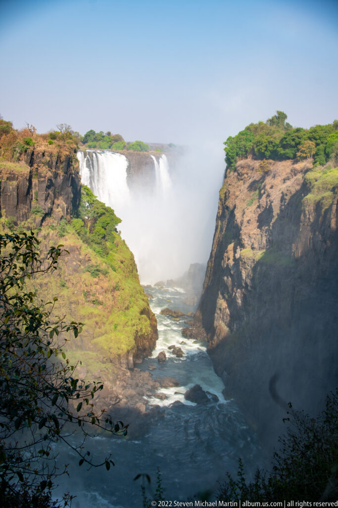 Broad Victoria Falls cascade beyond a deep basalt gorge with river water below and green vegetation on both rims.