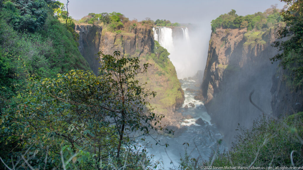 Forested viewpoint over the narrow basalt gorge below Victoria Falls, with a distant waterfall segment and mist rising between cliffs.