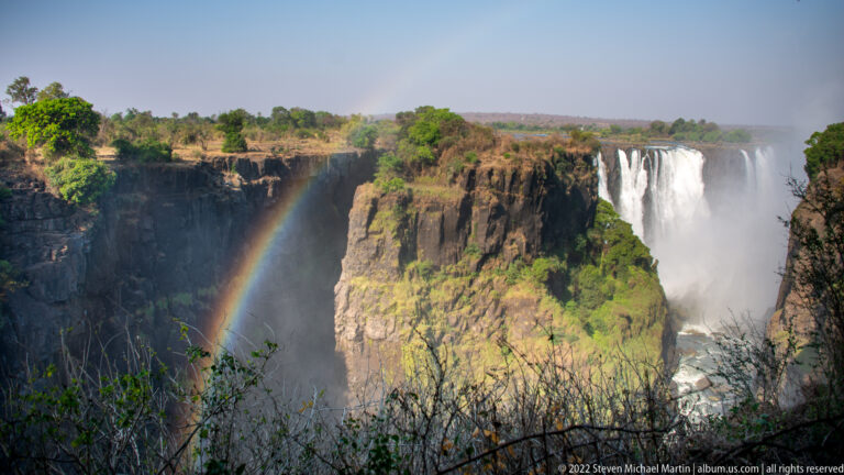 Rainbow rising from the mist-filled gorge beside a broad section of Victoria Falls, with sunlit basalt cliffs and vegetation.