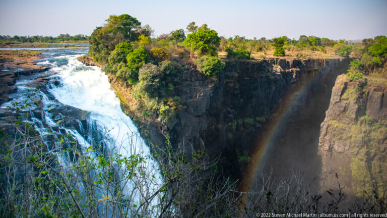 Close oblique view of water pouring over Victoria Falls beside a basalt cliff, with a narrow rainbow in the mist below.