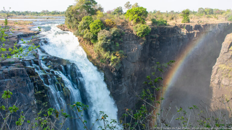 Broad white cascade at Victoria Falls dropping over dark rock, with a rainbow forming in spray beside the opposite cliff.